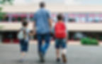 Padre caminando con sus dos hijos hacia la entrada de un colegio, llevando mochilas y comenzando su primer día en una nueva escuela en el extranjero.